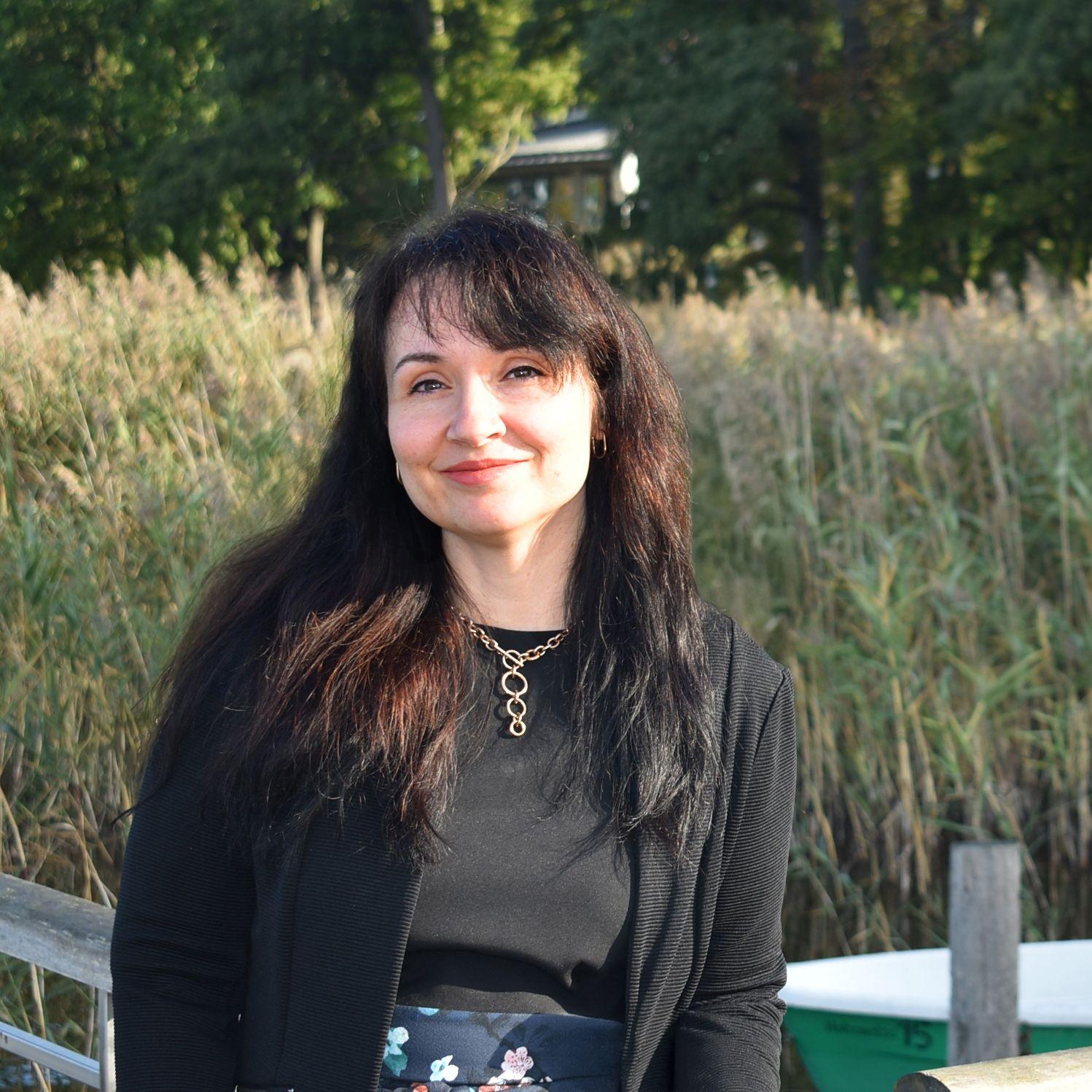 Portrait der Kaufmännischen Leiterin Sylvia Hofmeister. Frau Hofmeister hat dunkle lange Haare mit Pony, ein dunkles Shirt & einen dunkle Jacke darüber sowie eine goldene Kette. Im Hintergrund See und ein Steg nach der Rehaklinik Hohenelse.